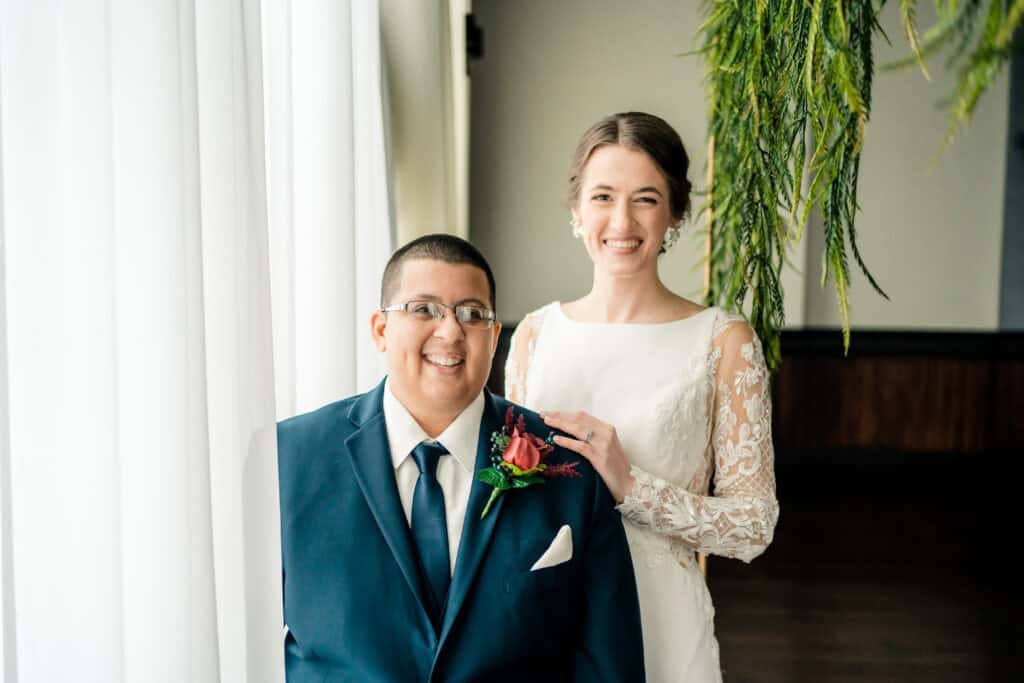 A couple, dressed in wedding attire, poses indoors by a window. The groom is in a dark suit with a red boutonnière, and the bride is in a white dress with lace sleeves, smiling with her hand on his shoulder.