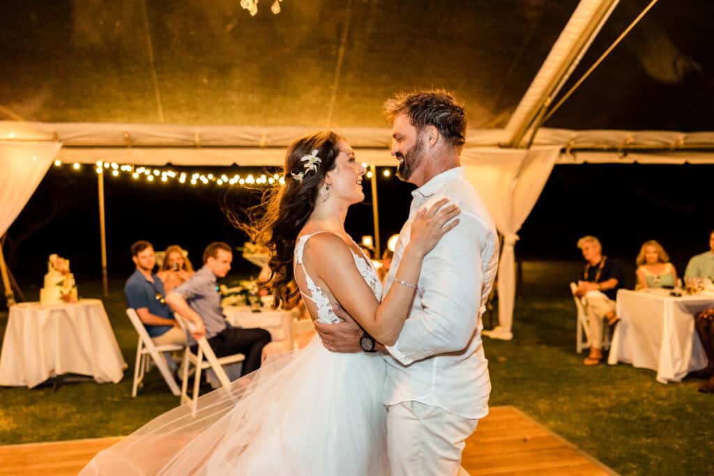 A bride and groom share a first dance under a tent with string lights, while guests seated at tables look on.