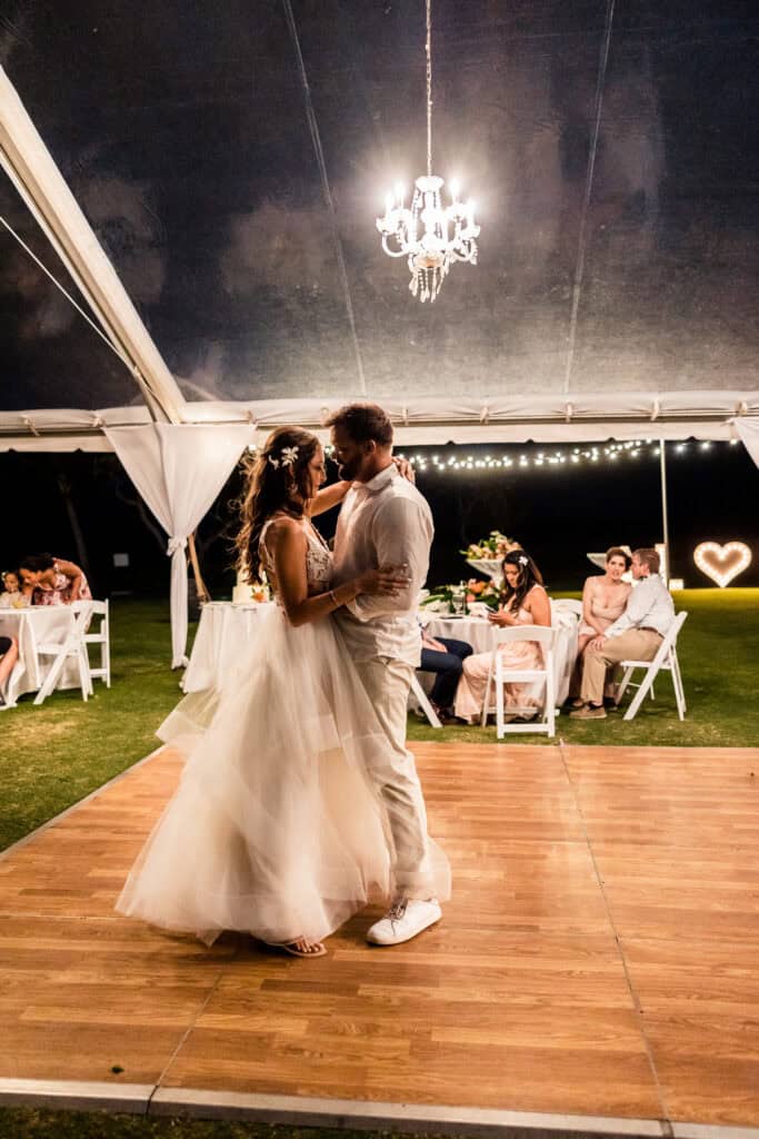 A bride and groom share their first dance on an outdoor dance floor under a chandelier as guests seated at tables watch.