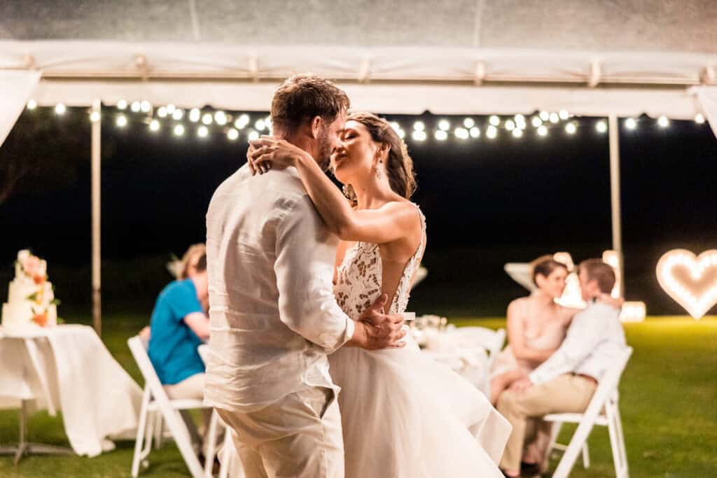 A bride and groom share a dance under string lights at an outdoor wedding reception, while guests sit at tables in the background.