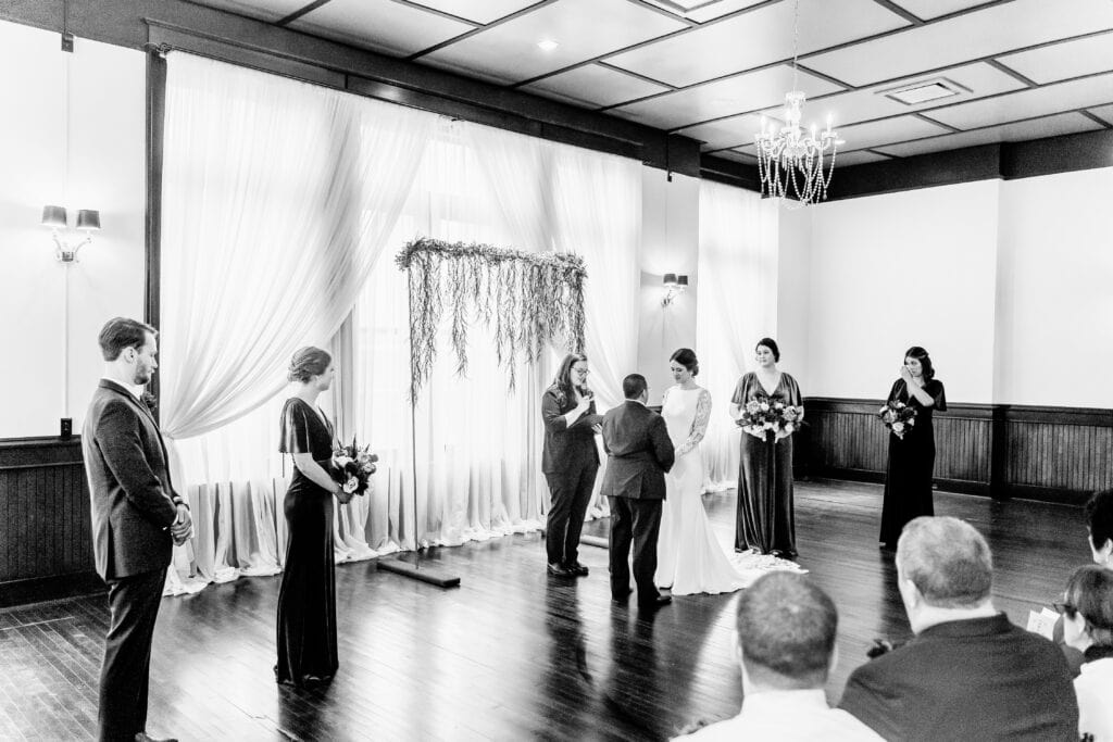 A black-and-white photo of a wedding ceremony. The couple stands in front of an officiant under a decorated arch, with the bridal party nearby. Guests are seated, watching the ceremony.