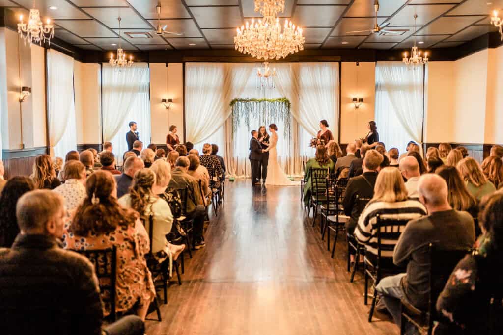 A wedding ceremony in a decorated hall with guests seated on both sides of the aisle, the couple stands at the altar exchanging vows under a floral arch.
