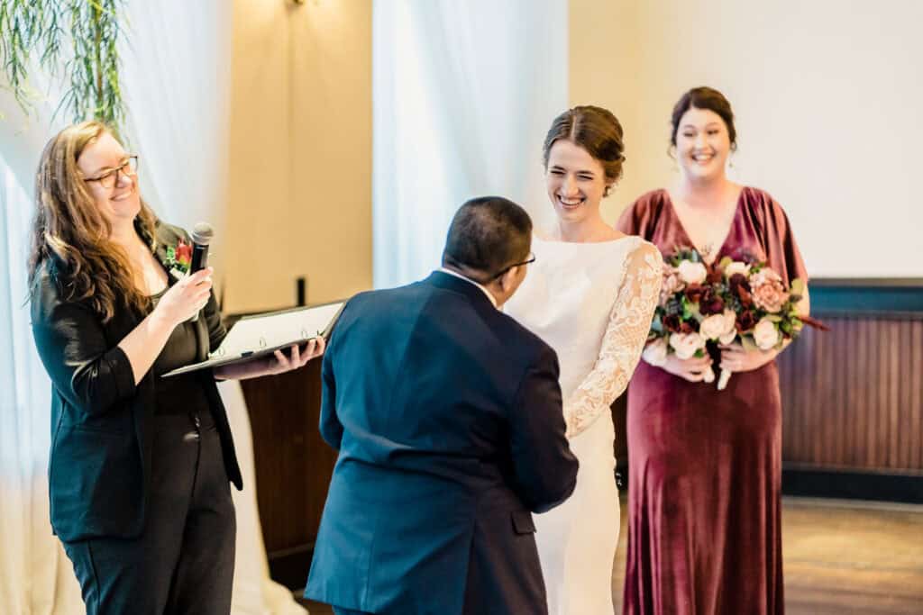 A wedding ceremony with a couple exchanging vows, officiant holding a microphone and ceremony book, and a bridesmaid holding a bouquet in the background.