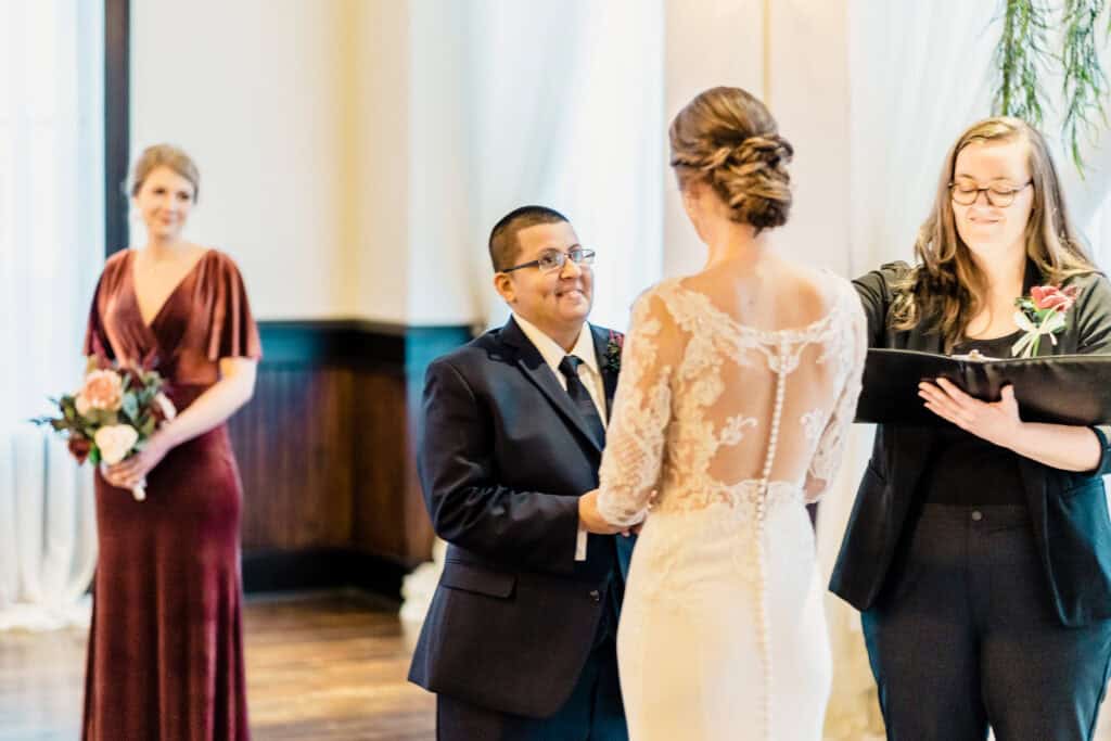 A couple in formal attire exchanges vows in front of an officiant, while a bridesmaid in a maroon dress, holding a bouquet, looks on.