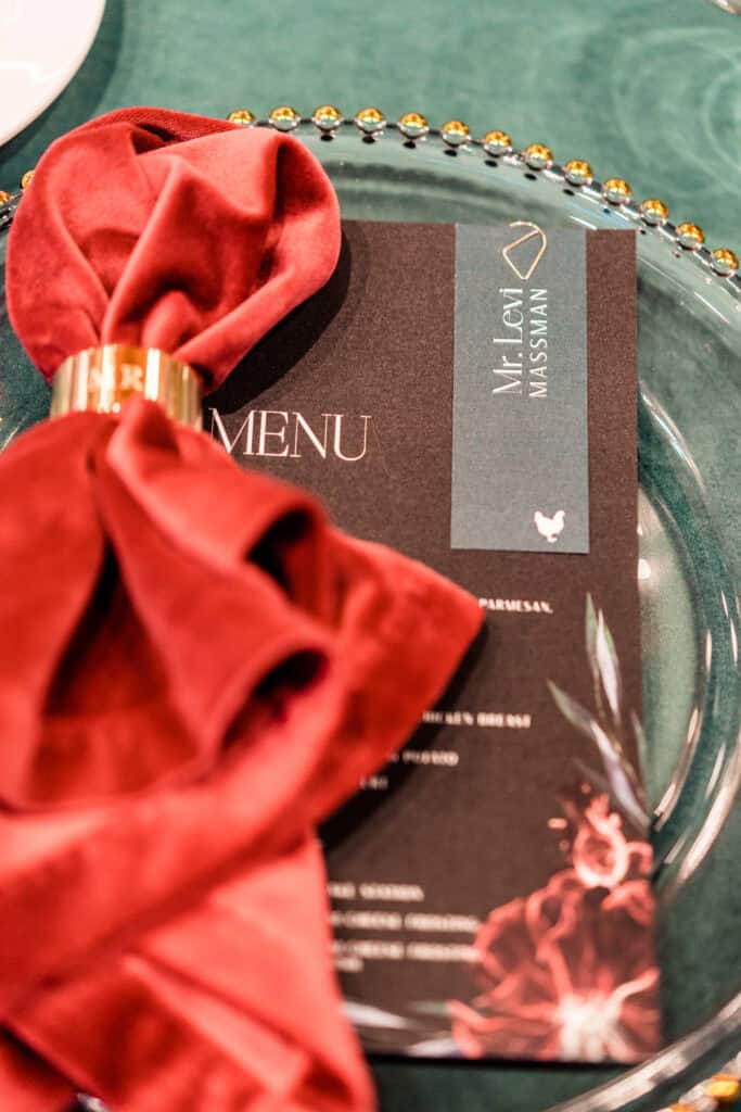 A dinner menu card with the name "Mr. Lea Nasswan" placed on a clear glass plate and a red fabric napkin held by a golden napkin ring. The tablecloth is green.