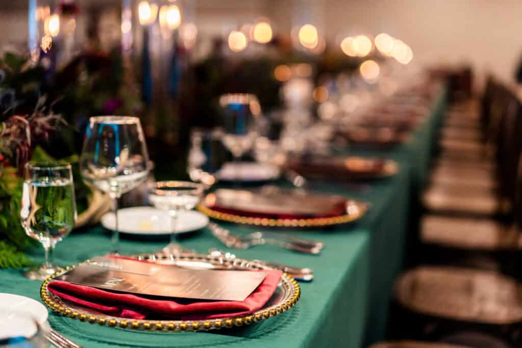 A long dining table is set for a formal event with plates, glasses, cutlery, and menus placed on a green tablecloth, illuminated by candlelight.