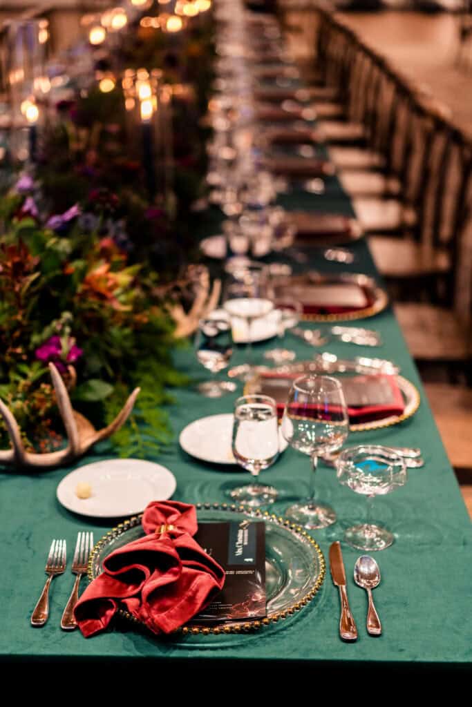 A long, elegant dining table is set with green tablecloth, silver cutlery, glassware, and decorated with flowers. A red napkin and a small black book are placed on each plate.