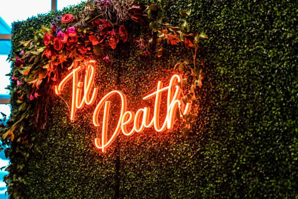 Neon sign displaying “Til Death” mounted on a green foliage wall with surrounding red flowers.
