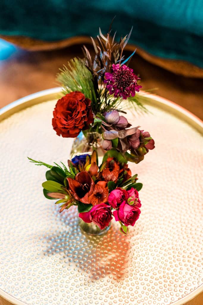 Two small floral arrangements with various colorful flowers and greenery in clear glass vases on a textured, round, beige table.