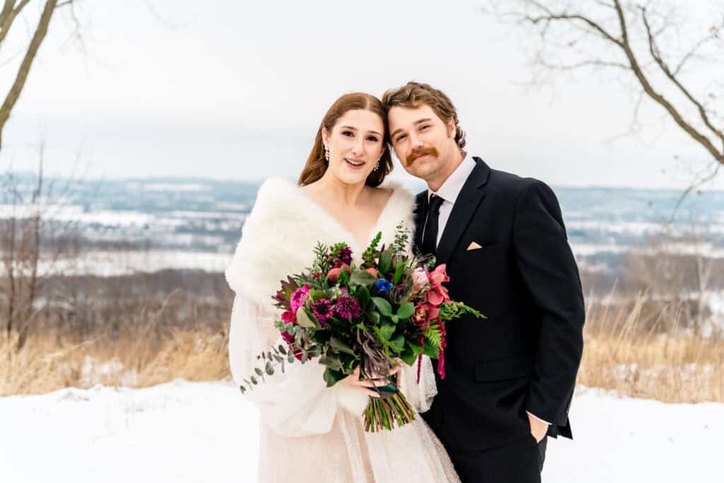 A couple stands together outdoors in winter, with snow on the ground. The woman holds a vibrant bouquet and wears a white shawl. The man wears a black suit and tie. Both are smiling.