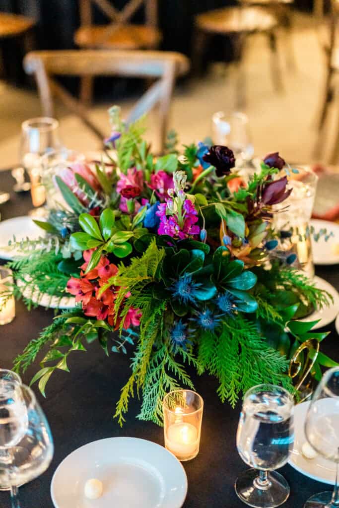 A table centerpiece with a vibrant floral arrangement surrounded by lit candles, glassware, and plates on a black tablecloth.