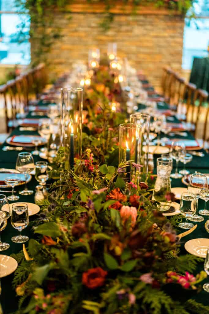 A long, elegantly set dining table with floral arrangements and lit candles running down the center, surrounded by neatly arranged plates, glasses, and silverware.