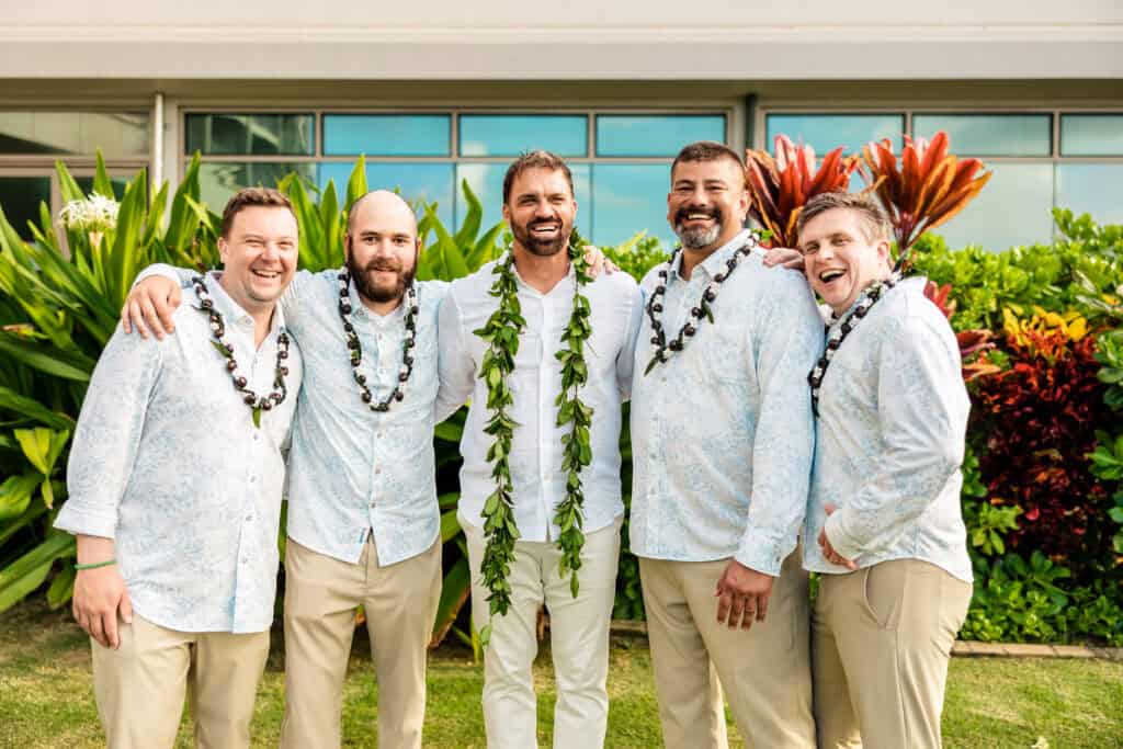 Five men wearing white shirts and beige pants, with one in the center adorned with a lei, standing outdoors in front of lush greenery, smiling, and posing for a photo.