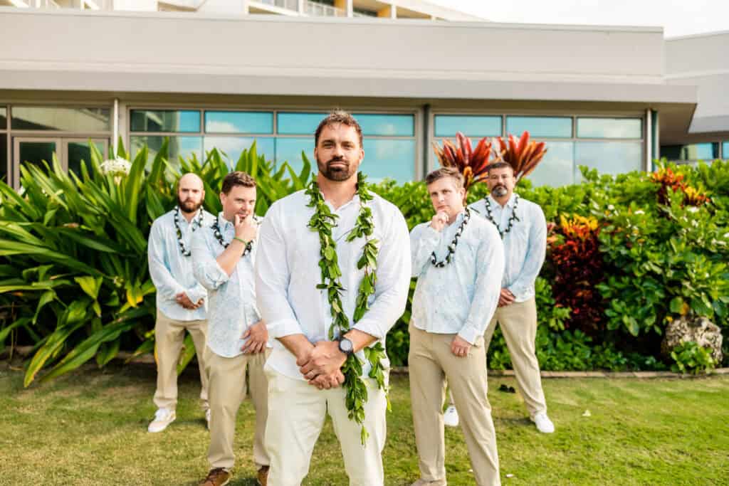 A groom stands in the foreground wearing a leafy lei, while four groomsmen in matching attire pose behind him in a landscaped garden setting.