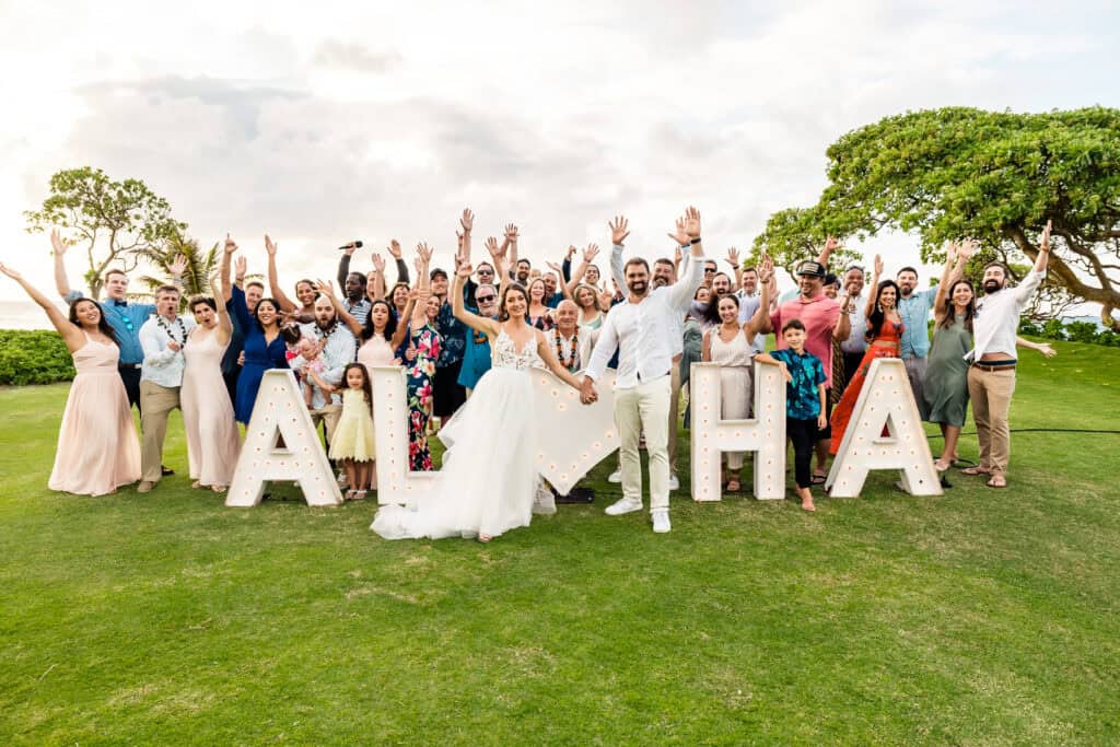 A large group of people, including the bride and groom, gather outdoors in front of illuminated "ALOHA" letters, with everyone smiling and raising their hands.