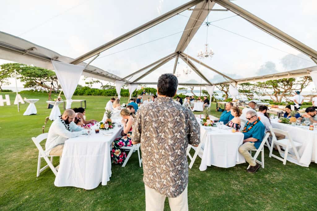 A person addresses a gathering at a wedding reception under a transparent tent, with guests seated at tables on a lawn.