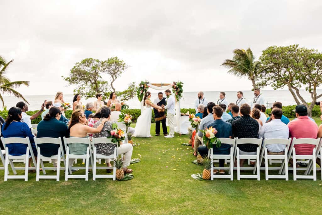 A wedding ceremony taking place outdoors by the ocean with guests seated in rows of white chairs. The couple is standing under an altar decorated with white fabric and greenery.