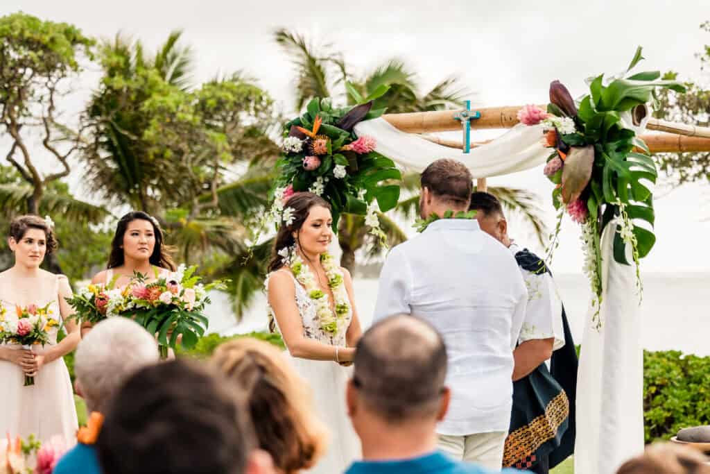 A couple stands under a floral arch during an outdoor wedding ceremony, exchanging vows. Bridesmaids holding bouquets observe from the side, and guests look on from their seats against a backdrop of lush greenery.
