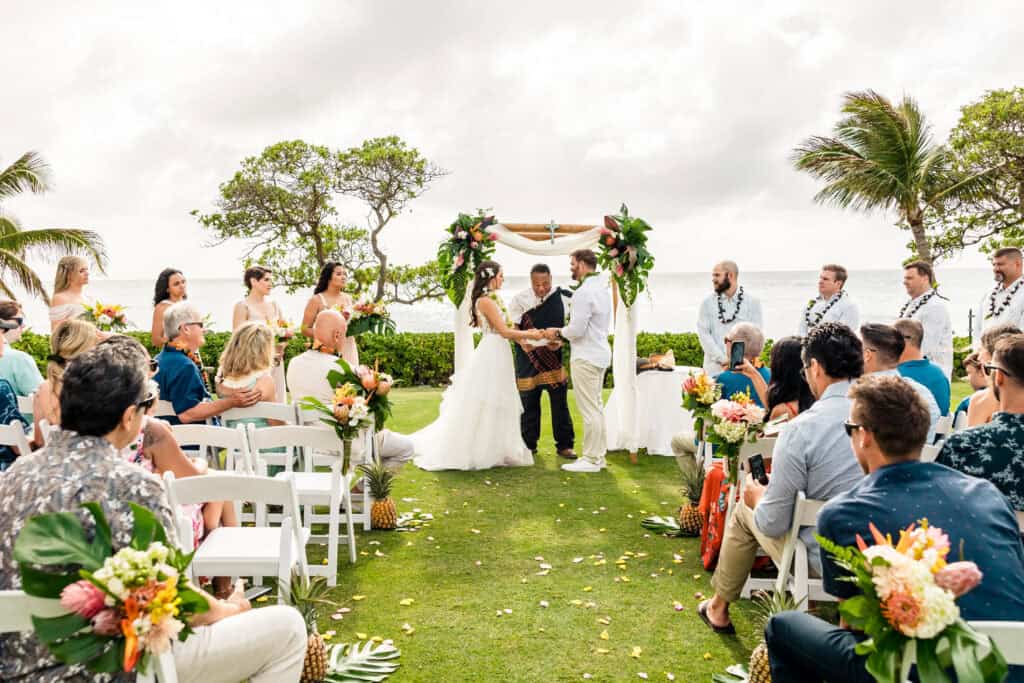 A couple stands at an altar adorned with flowers, exchanging vows in an outdoor wedding ceremony by the ocean. Guests are seated on both sides, with floral arrangements along the aisle.