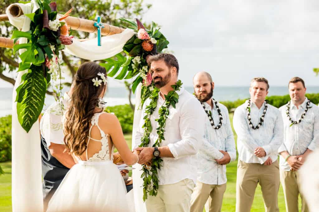 A couple exchanges vows under a decorated arbor at an outdoor wedding ceremony by the sea, with three men standing as groomsmen and lush greenery in the background.