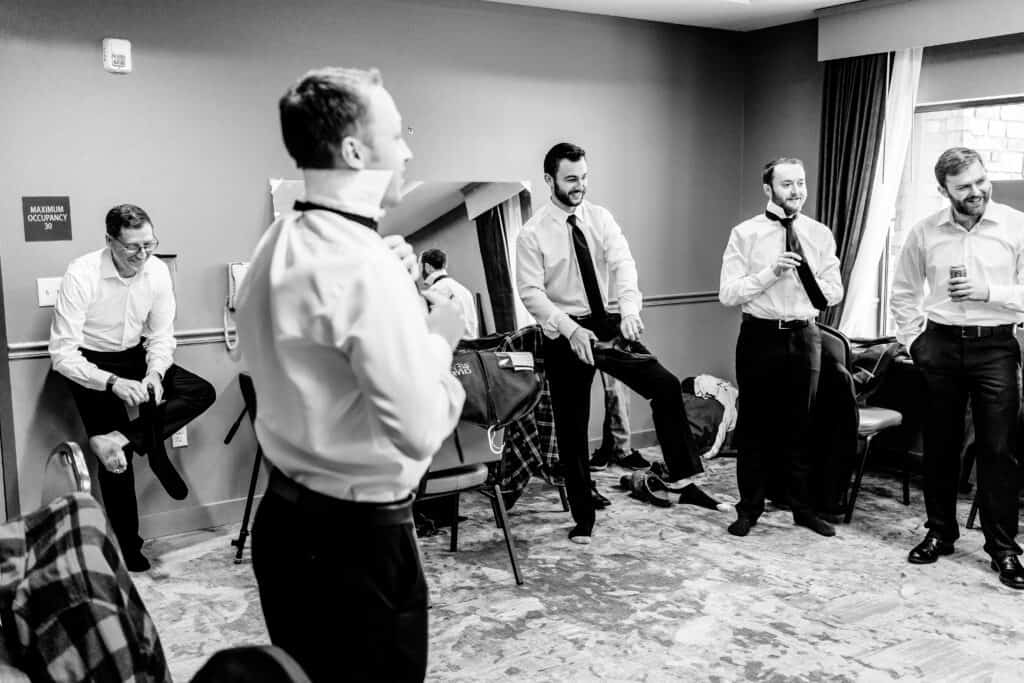 A group of men in dress shirts and pants are preparing for an event in a room. Some are adjusting their neckties, while others are standing or sitting and talking.