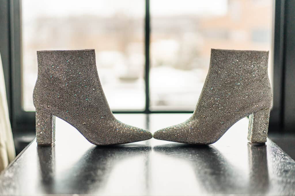 A pair of sparkly, silver high-heeled ankle boots are displayed side by side with a window in the background.