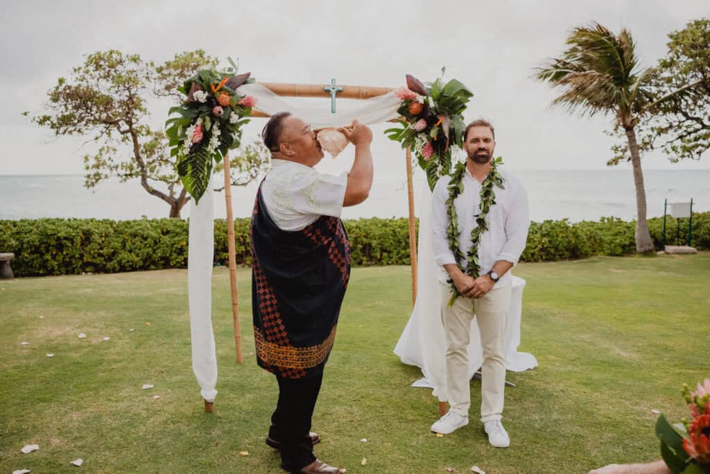 A person blows into a conch shell near a floral decorated arch on a grassy area by the sea. Another person dressed in white with a green lei stands next to the arch, with the ocean and trees in the background.