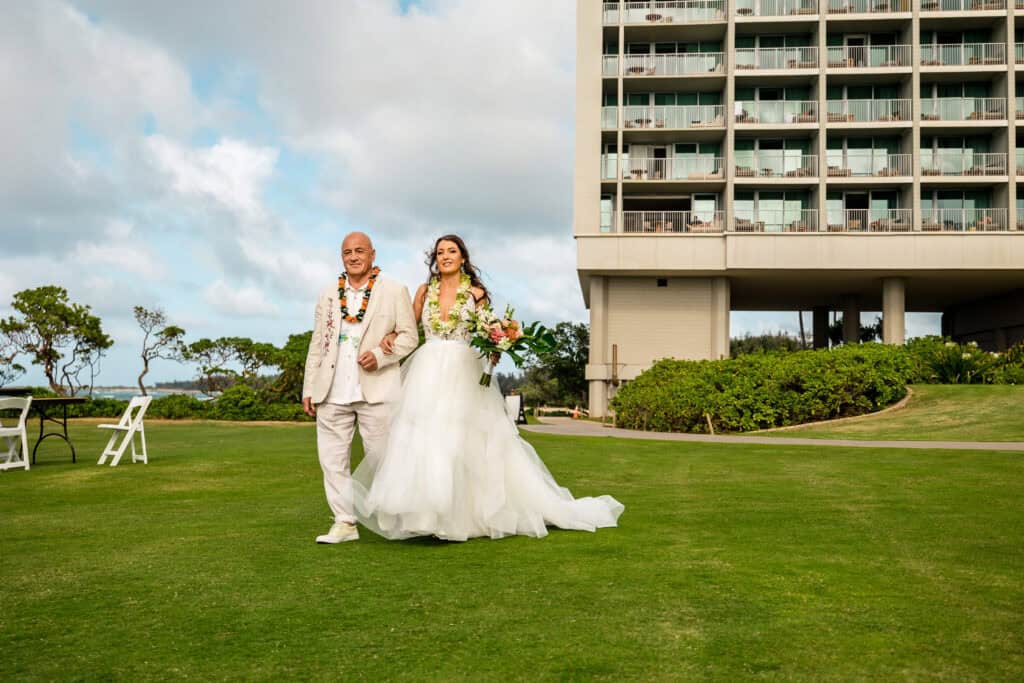 A bride in a white dress walks arm-in-arm with a man in a light-colored suit on a grassy area next to a tall building, with trees and a cloudy sky in the background.