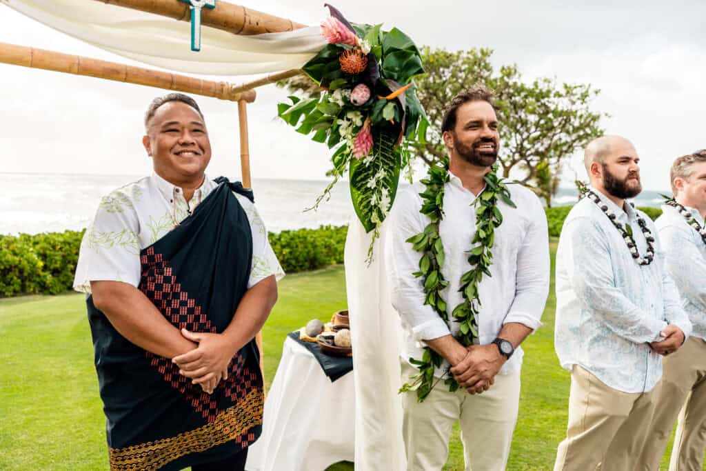 Two men in ceremonial attire stand on a grassy area by the ocean, beneath a woven arch adorned with tropical flowers and leaves. A third man stands nearby, wearing a lei and white shirt.