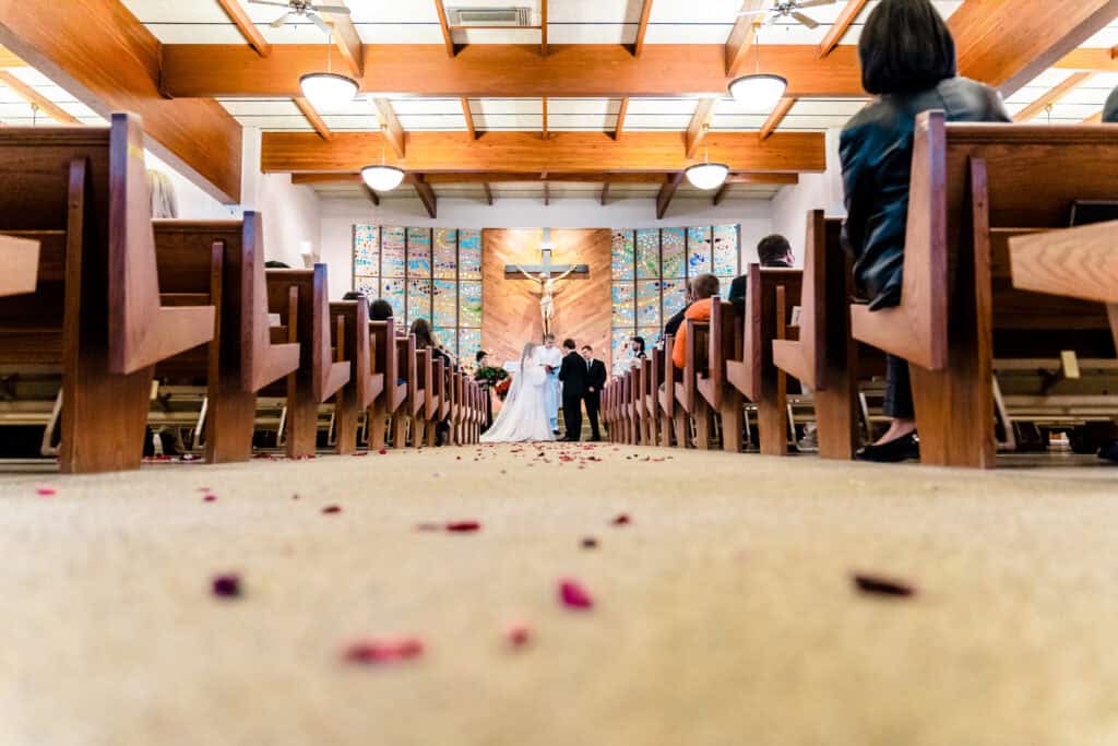 A couple stands at the altar in a church, seen from the aisle with scattered rose petals, guests seated on both sides.