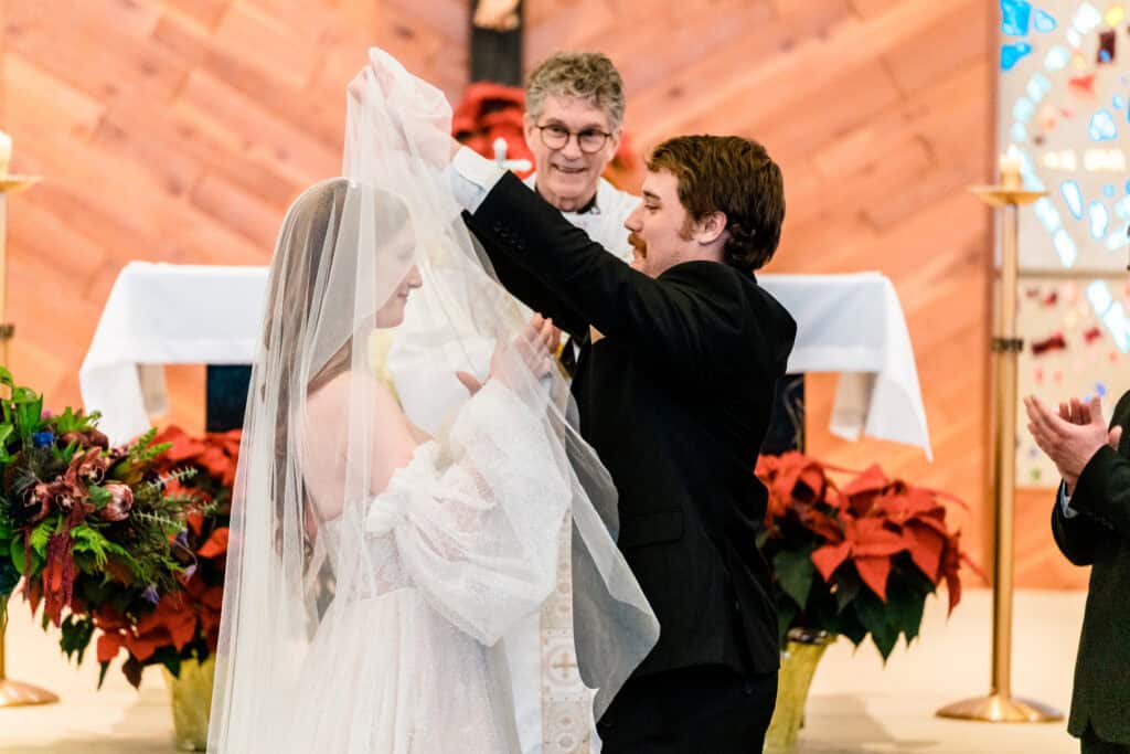 A groom lifts the bride's veil during a wedding ceremony as a smiling officiant stands behind them in a church adorned with flowers.