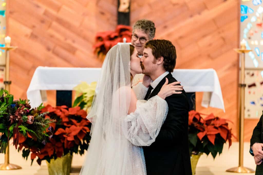 A bride and groom share a kiss in front of an altar during their wedding ceremony, with an officiant and floral arrangements in the background.