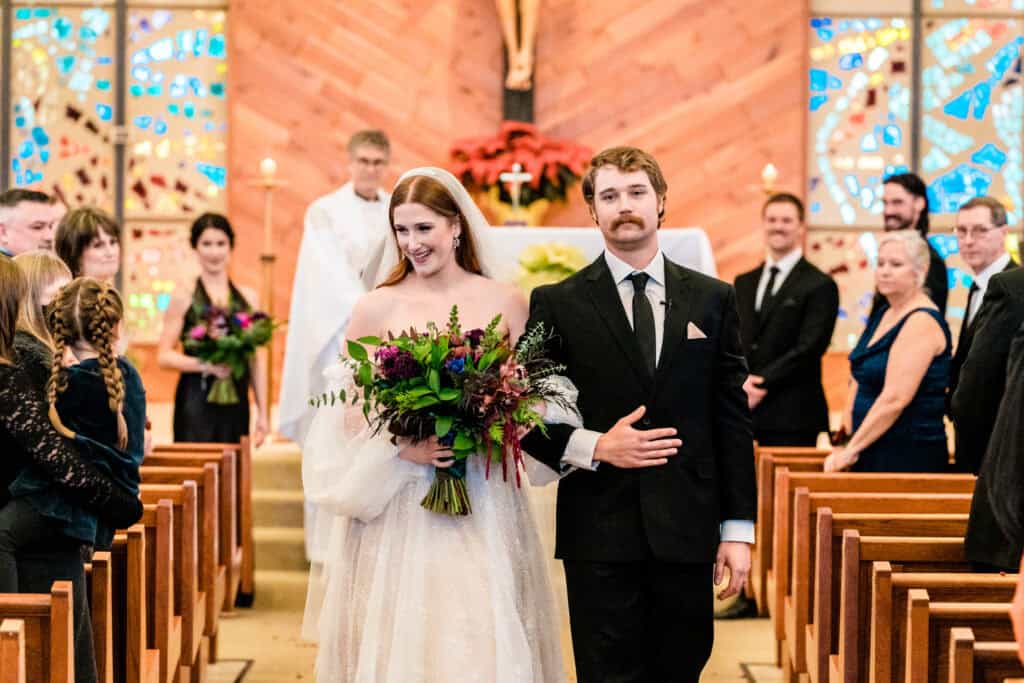 A bride and groom walk down the aisle together in a church, smiling and holding a bouquet. The groom has his arm around the bride. Guests in formal attire look on and smile.
