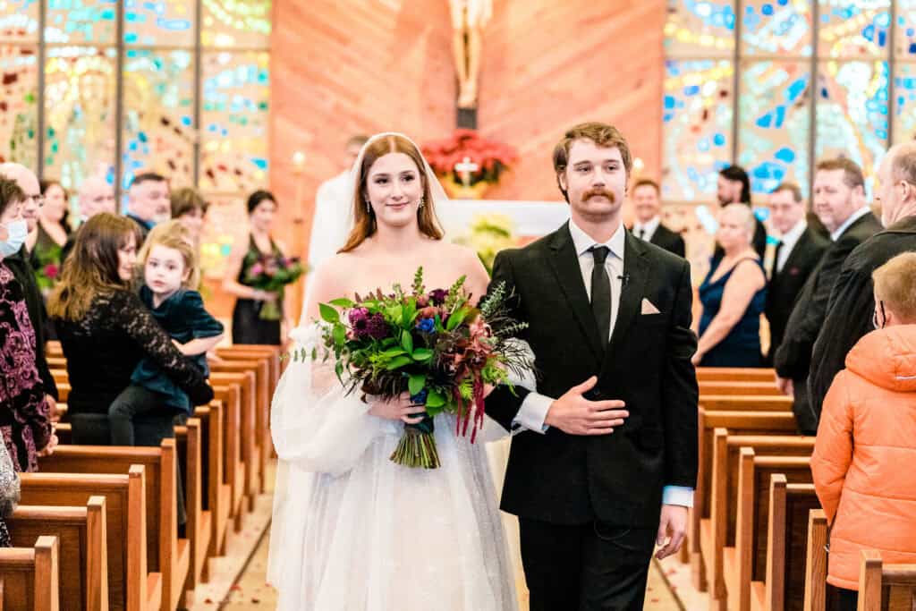 A bride and groom walk down the aisle together in a church filled with guests. The bride is holding a bouquet of flowers, and the groom has his arm around her.