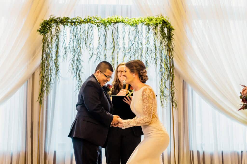 A couple stands facing each other at their wedding ceremony, holding hands. An officiant in the background presides over the event. An arch decorated with greenery is behind them.