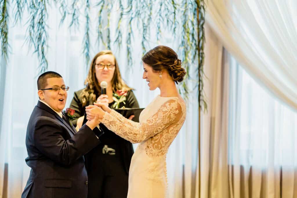 A couple holds hands during a wedding ceremony with an officiant standing behind them. The bride wears a long-sleeved lace dress and the groom is in a dark suit. They appear to be smiling and joyful.