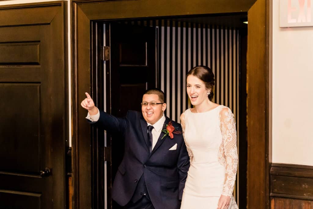 A couple in formal attire is entering a room through a doorway. Both are smiling; the person on the left is pointing forward. The person on the right is wearing a white dress with lace sleeves.