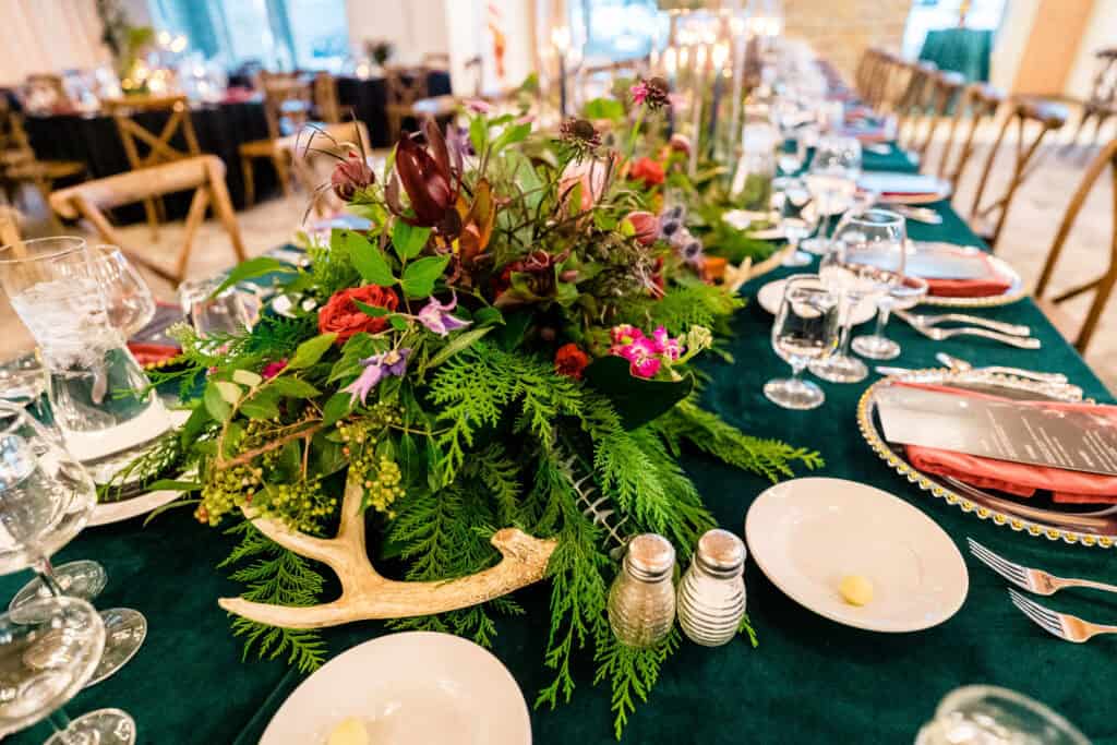 A long table set for a meal, decorated with green foliage, pink and purple flowers, and antler centerpiece. Plates, glasses, salt and pepper shakers are neatly arranged with cutlery.