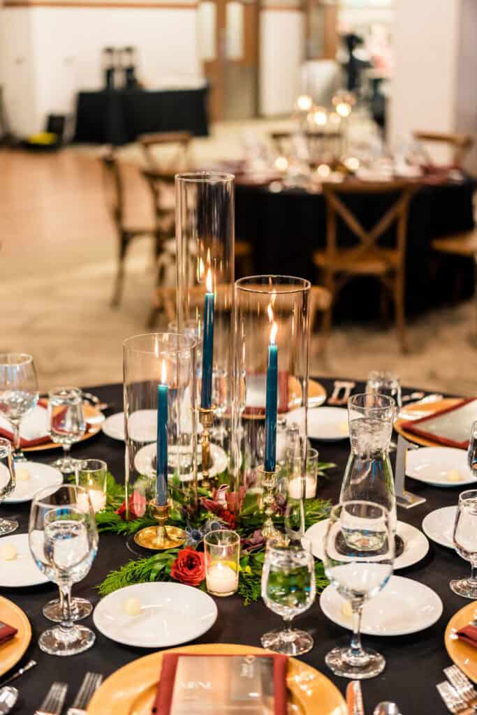 A round, elegantly set dining table featuring blue candles in tall glass holders, surrounded by glassware, plates, and a floral centerpiece. Chairs and another set table are visible in the background.