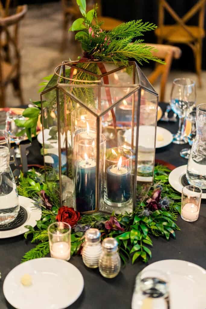 Elegant table setting featuring a centerpiece with candles inside a glass lantern, surrounded by greenery. Plates, utensils, glasses, and small candles are arranged around the centerpiece.