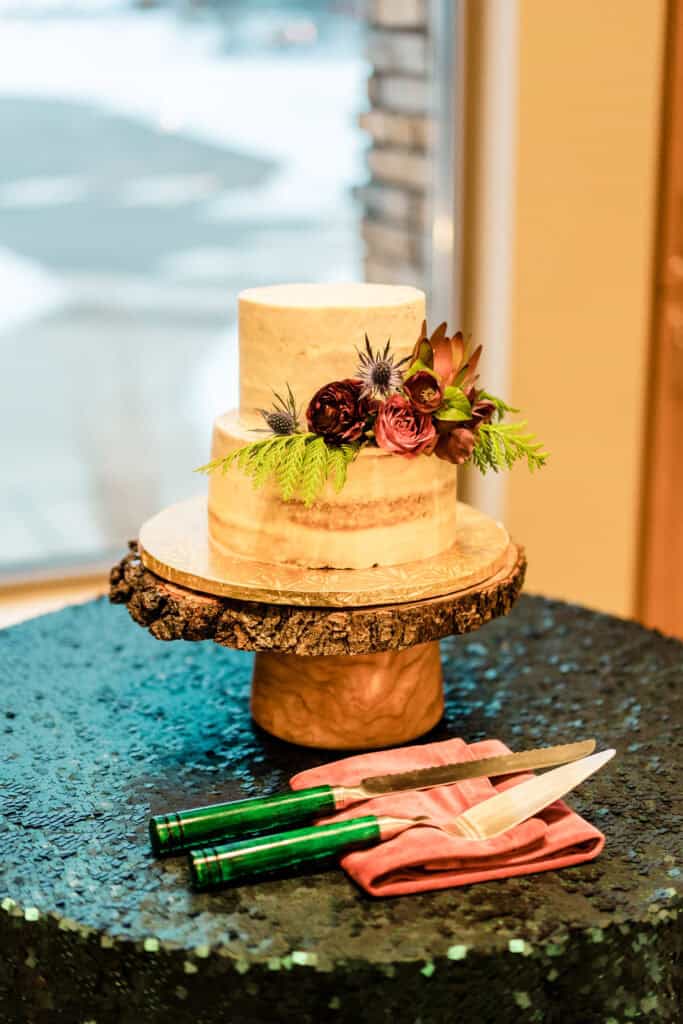 Two-tiered naked wedding cake adorned with flowers and greenery, placed on a wooden stand. Cake cutting knife and server rest on a pink napkin beside it.
