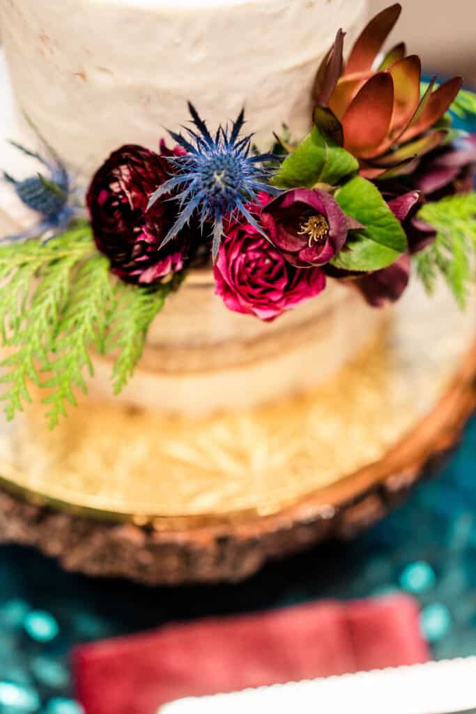 A close-up view of a decorated cake with a floral arrangement featuring red, blue, and brown flowers, greenery, and placed on a glass stand with a wooden base.