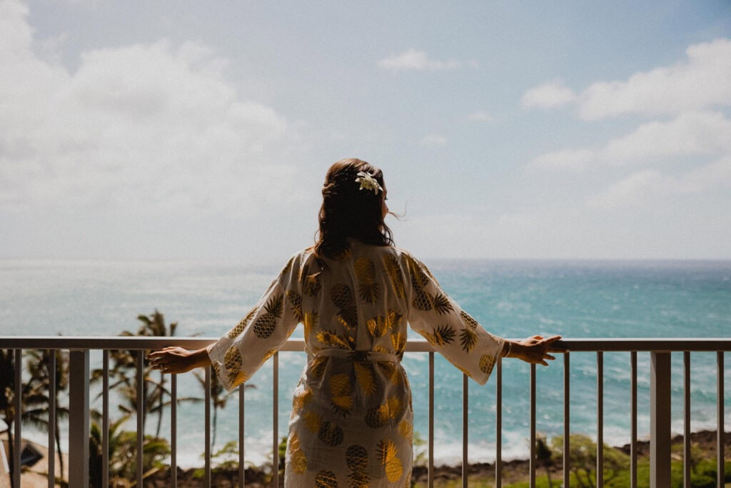 A person with a floral hair accessory stands on a balcony overlooking a scenic ocean view under a partly cloudy sky. They are wearing a robe with a pineapple pattern.