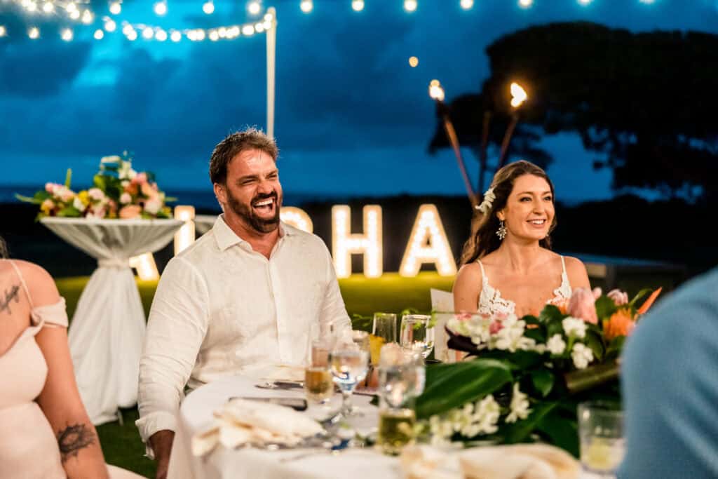 A man and a woman in formal attire sit at a dinner table outdoors at night, smiling. String lights and floral arrangements decorate the area, with illuminated letters in the background spelling "ALOHA.