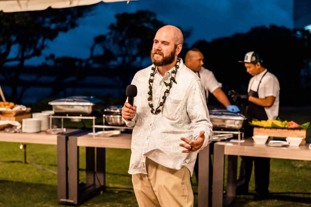 A man with a beard, wearing a white shirt and a lei, holds a microphone while speaking outdoors at night. Two catering staff members are working at a table behind him.