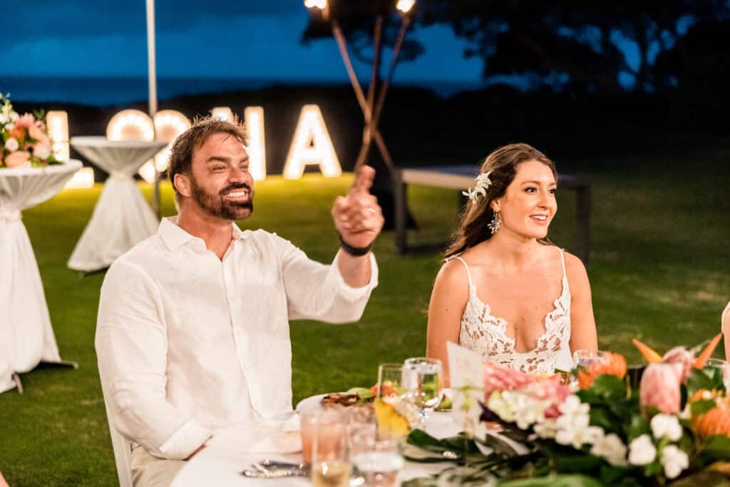 A man and a woman are seated together at an outdoor event during the evening. The man is gesturing with a smile, and the woman looks ahead. Illuminated "ALOHA" letters and floral decorations are in the background.