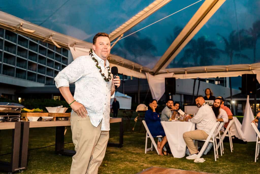 A man stands and speaks into a microphone at an outdoor event under a tent while seated guests listen.