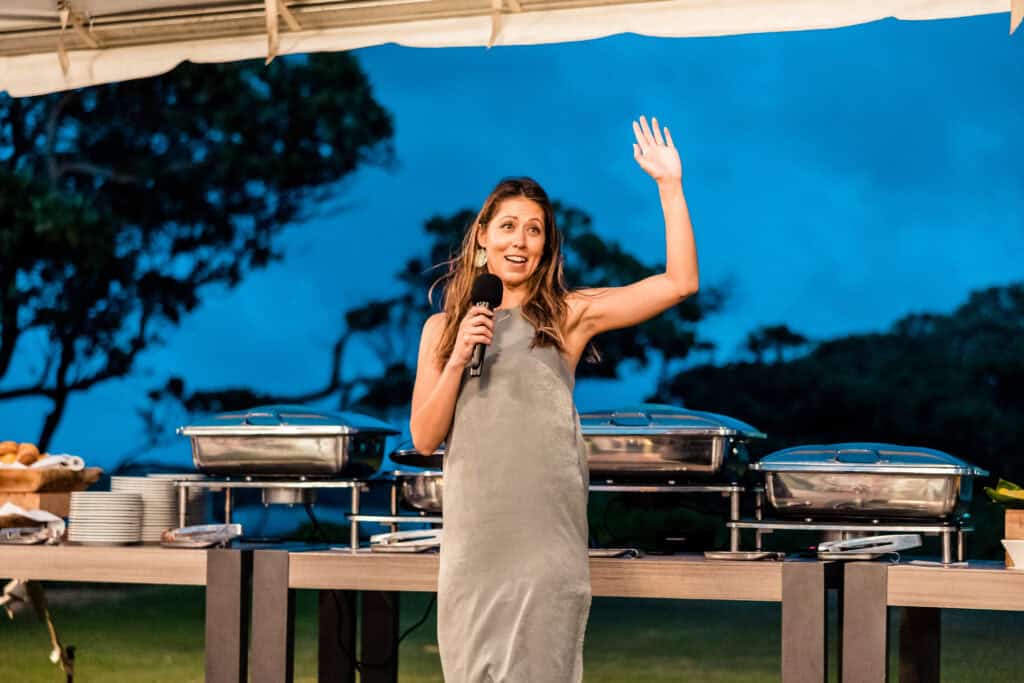 A person with long hair, wearing a gray dress, speaks into a microphone while raising their hand. They stand in front of a buffet table loaded with covered dishes, set outdoors in an evening setting.