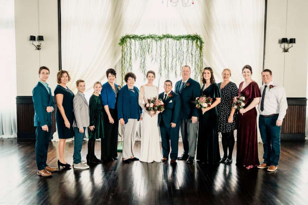 A wedding party with ten individuals dressed in formal attire standing on each side of the couple at the center, who are holding bouquets. The backdrop features white curtains and green foliage.