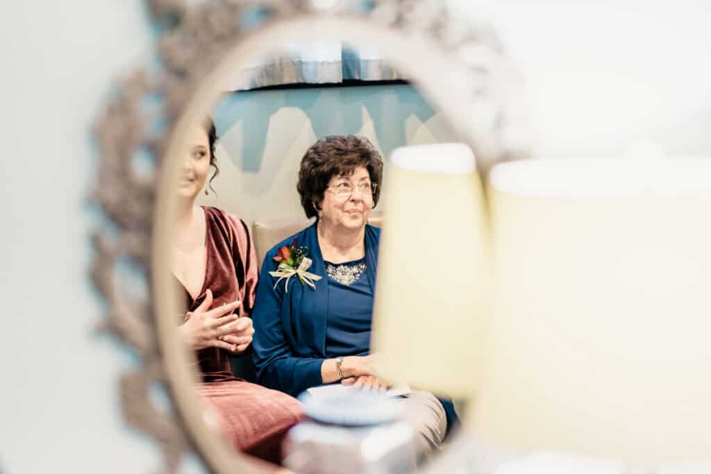 A mirror reflection shows two women seated; the older woman wears a blue top with a floral accessory, while the younger woman wears a maroon dress. A blurred lamp is in the foreground.